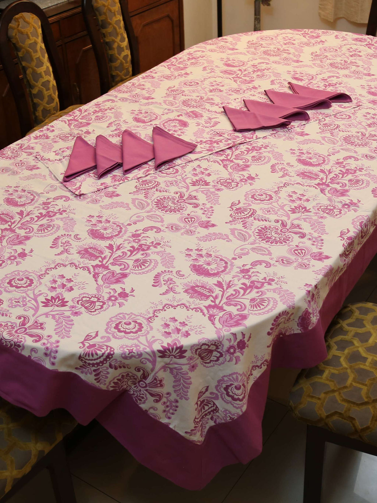 Off-white oval table cover set with a pink and purple paisley floral print, a solid mauve-pink border, and matching mauve triangle-folded napkins