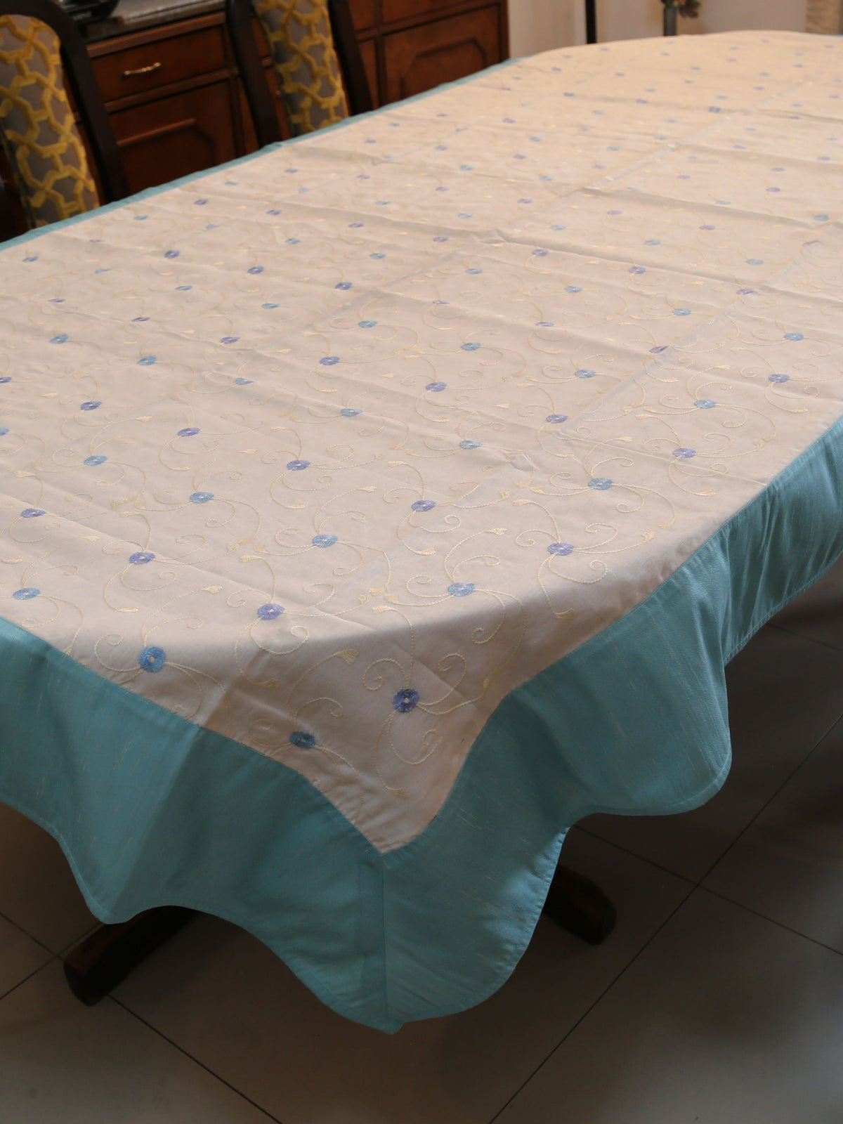 White oval table cover with small blue embroidered flower-and-vine dots and a solid aqua-teal border