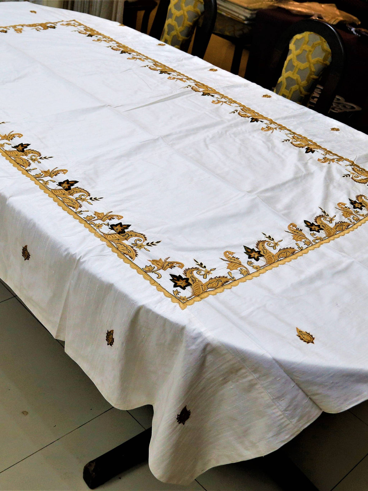 White table cover with a gold-brown embroidered paisley-and-vine corner border and scattered small leaf motifs