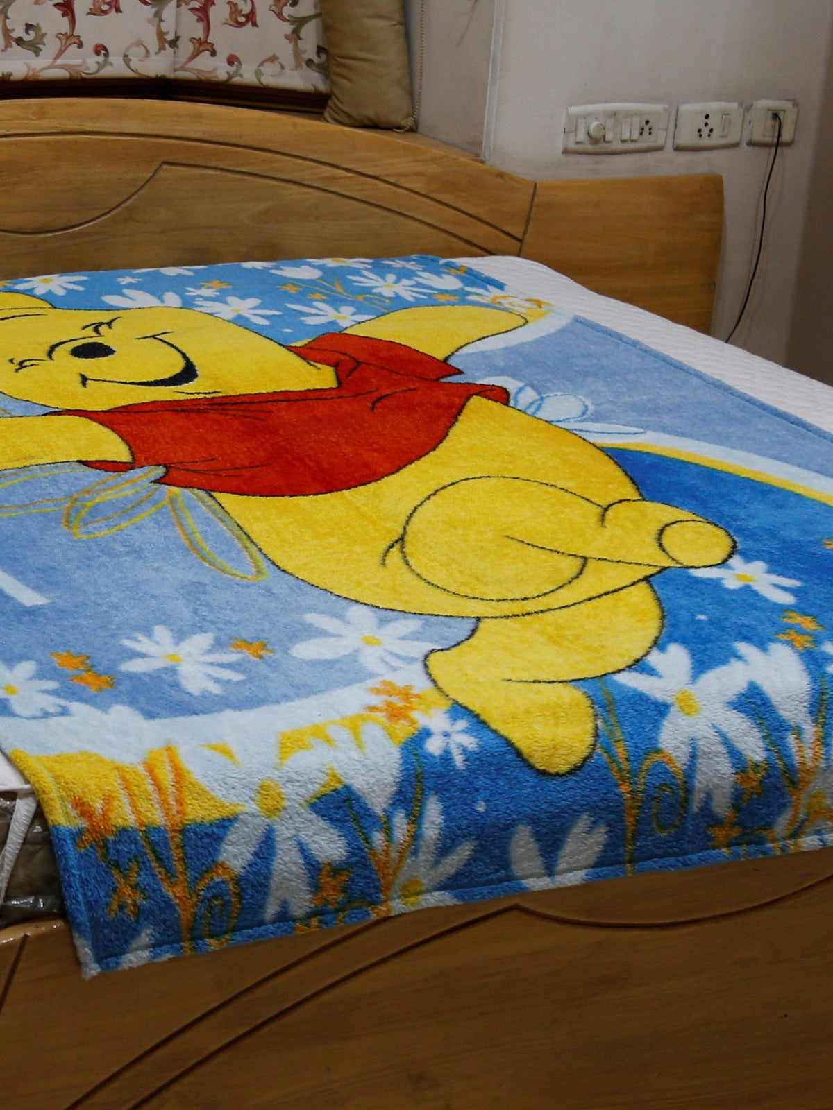 Blue plush blanket with a printed Winnie the Pooh design over a daisy-print border, draped over a bed