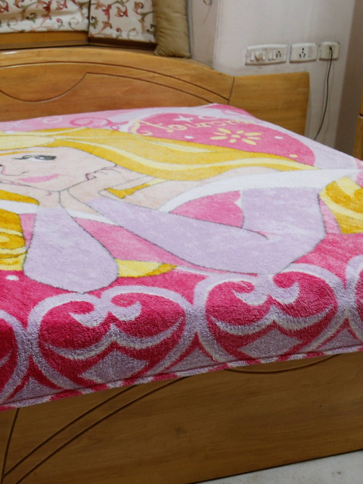 Pink and lilac blanket with a printed Disney Princess Aurora design and a scroll-heart border, draped over a bed