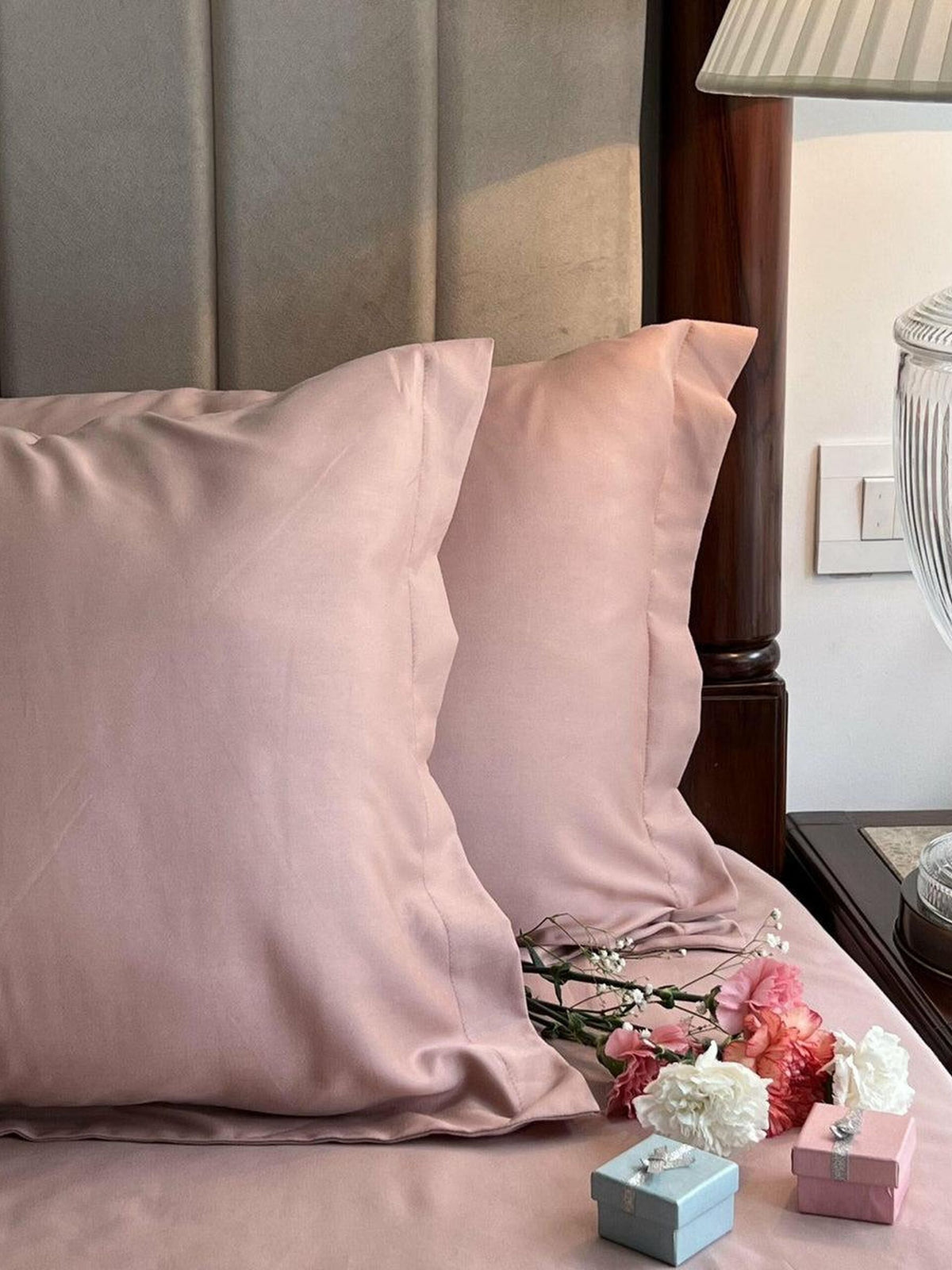 Dusty rose-pink satin pillow covers styled on a bed with dried baby's breath flowers, fresh carnations, and small gift boxes