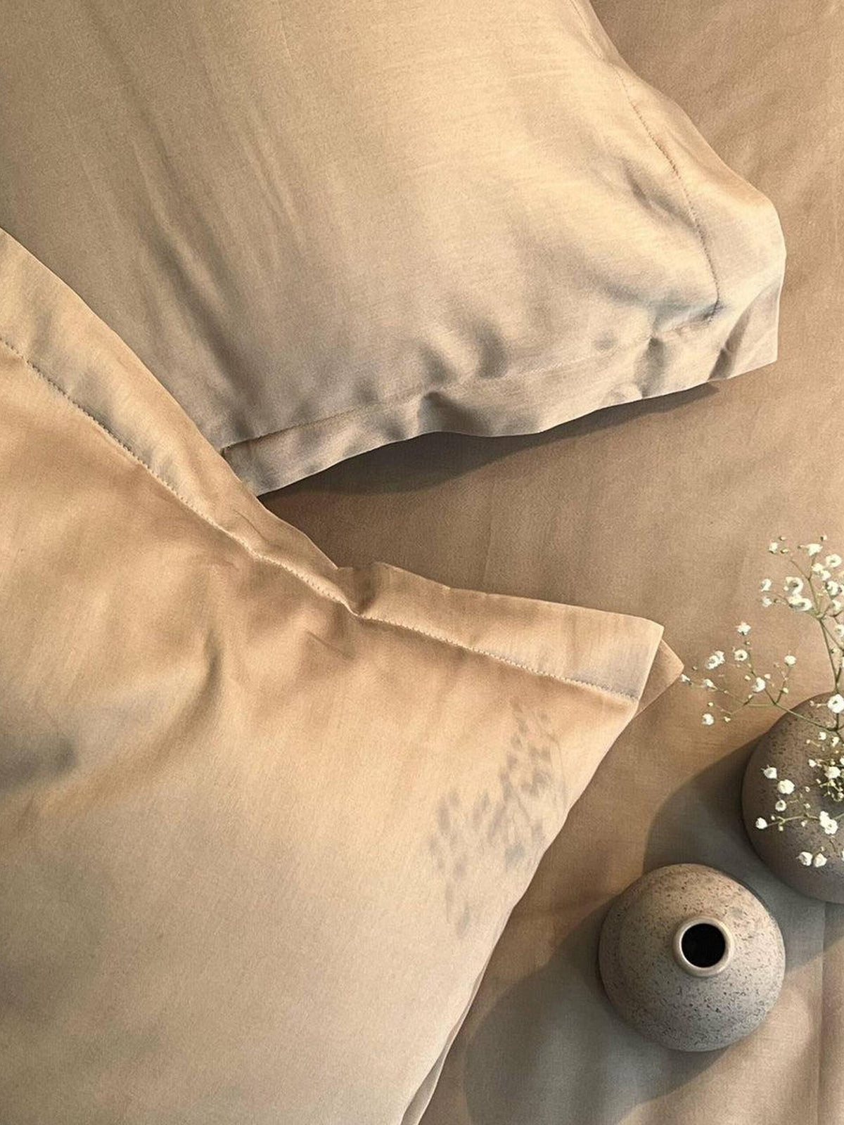 Taupe-sand satin bedsheet and pillow covers styled on a bed with a small vase of blue flowers and baby's breath