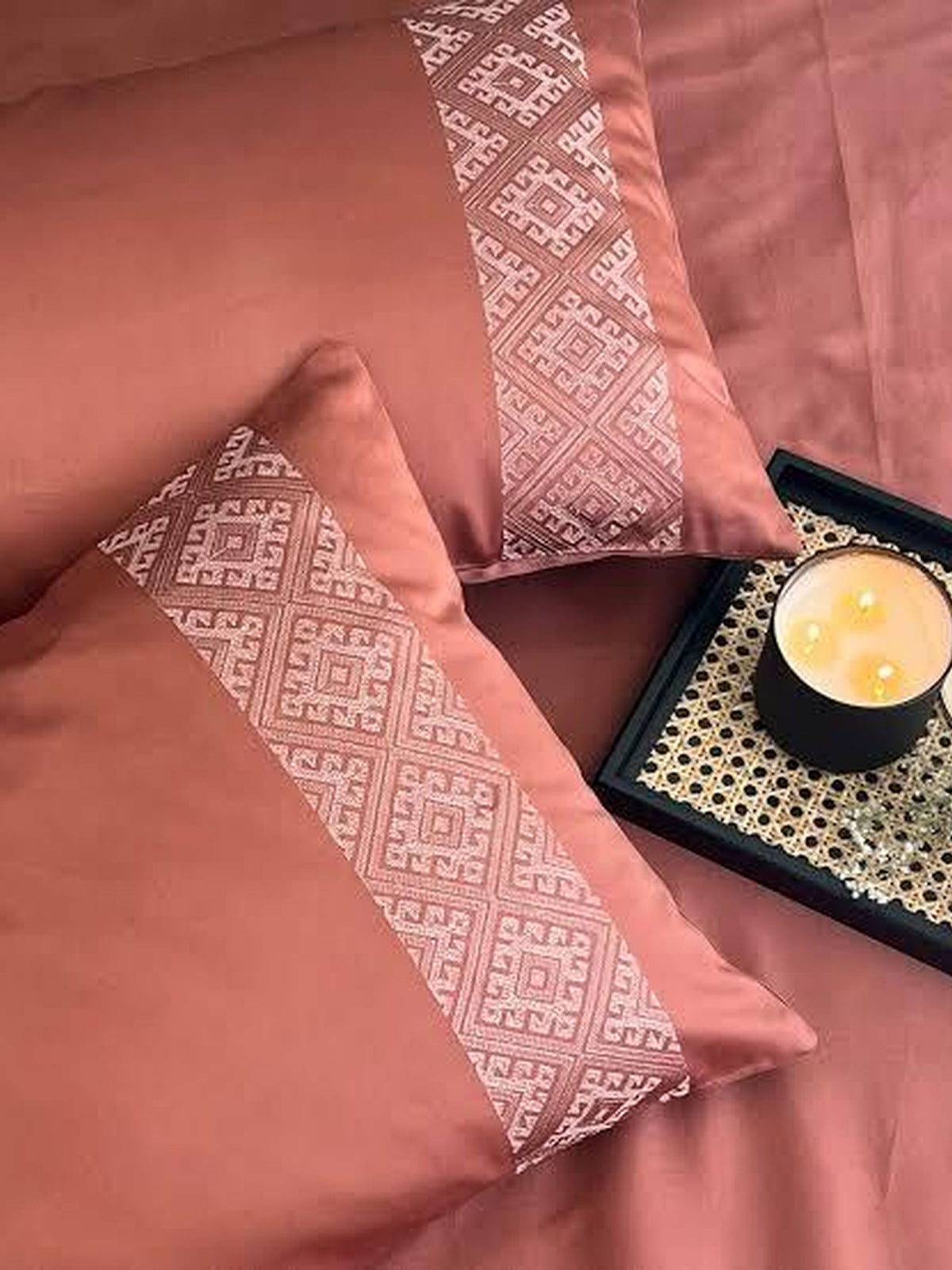 Terracotta bedsheet and pillow covers with a beige diamond-lattice embroidered band, styled on a bed with a lit candle on a cane tray
