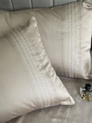Beige satin pillow covers with a dotted vertical-stripe embroidered band, styled on a bed with a perfume bottle