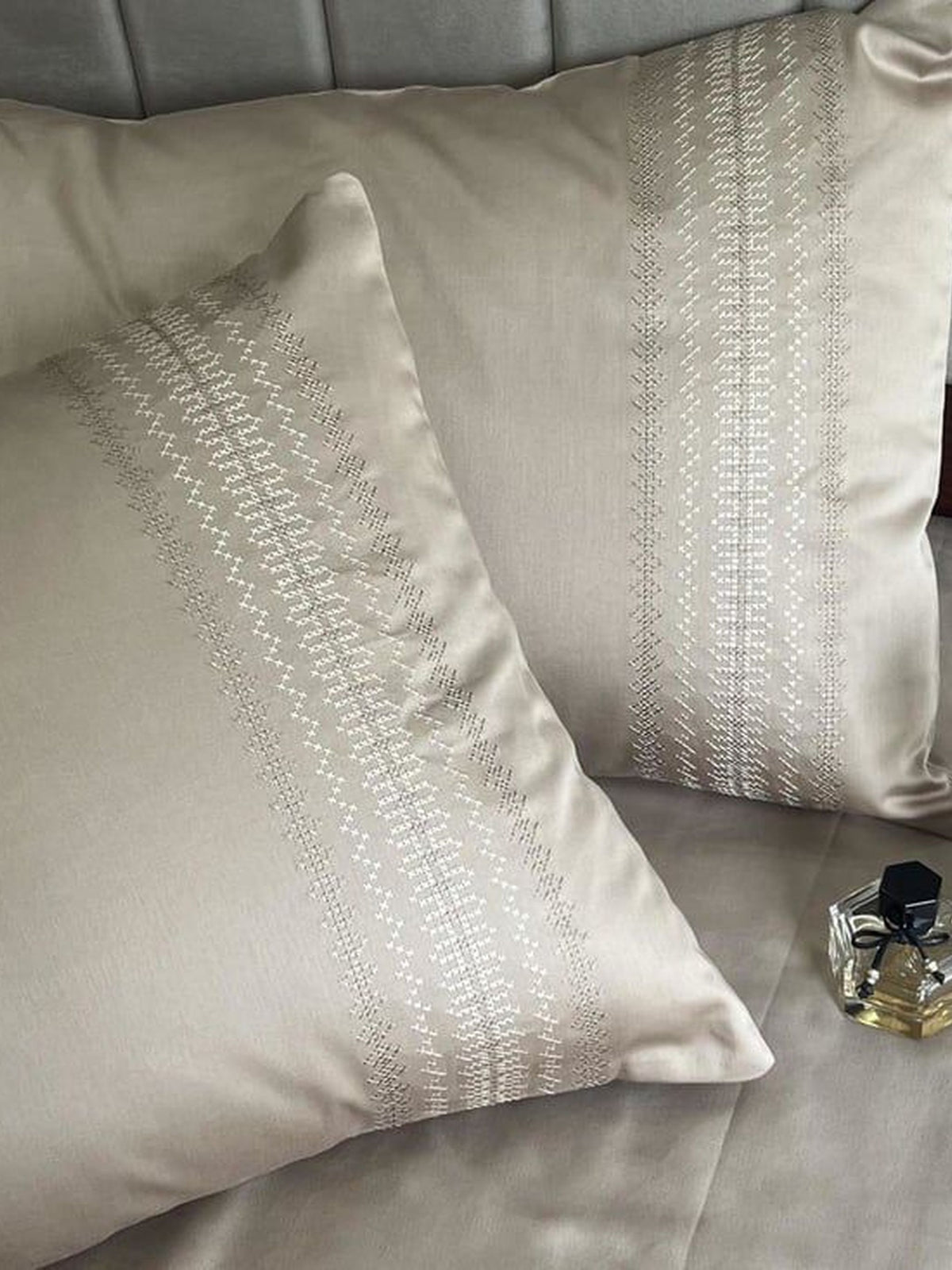 Beige satin pillow covers with a dotted vertical-stripe embroidered band, styled on a bed with a perfume bottle