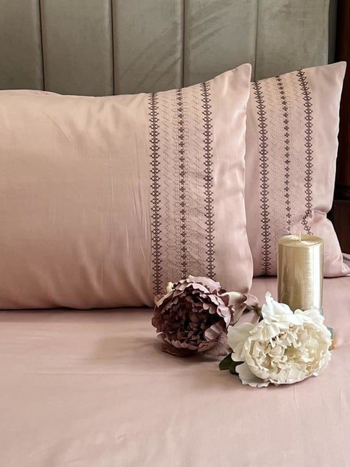 Blush-pink bedsheet and pillow covers with a brown cross-stitch striped embroidered band, styled on a bed with a candle and dried flowers