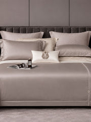 Taupe grey bedding with a fine embroidered border on a grey upholstered bed