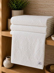 White cotton bath towel with an all-over floral damask pattern folded on a wooden bathroom shelf.