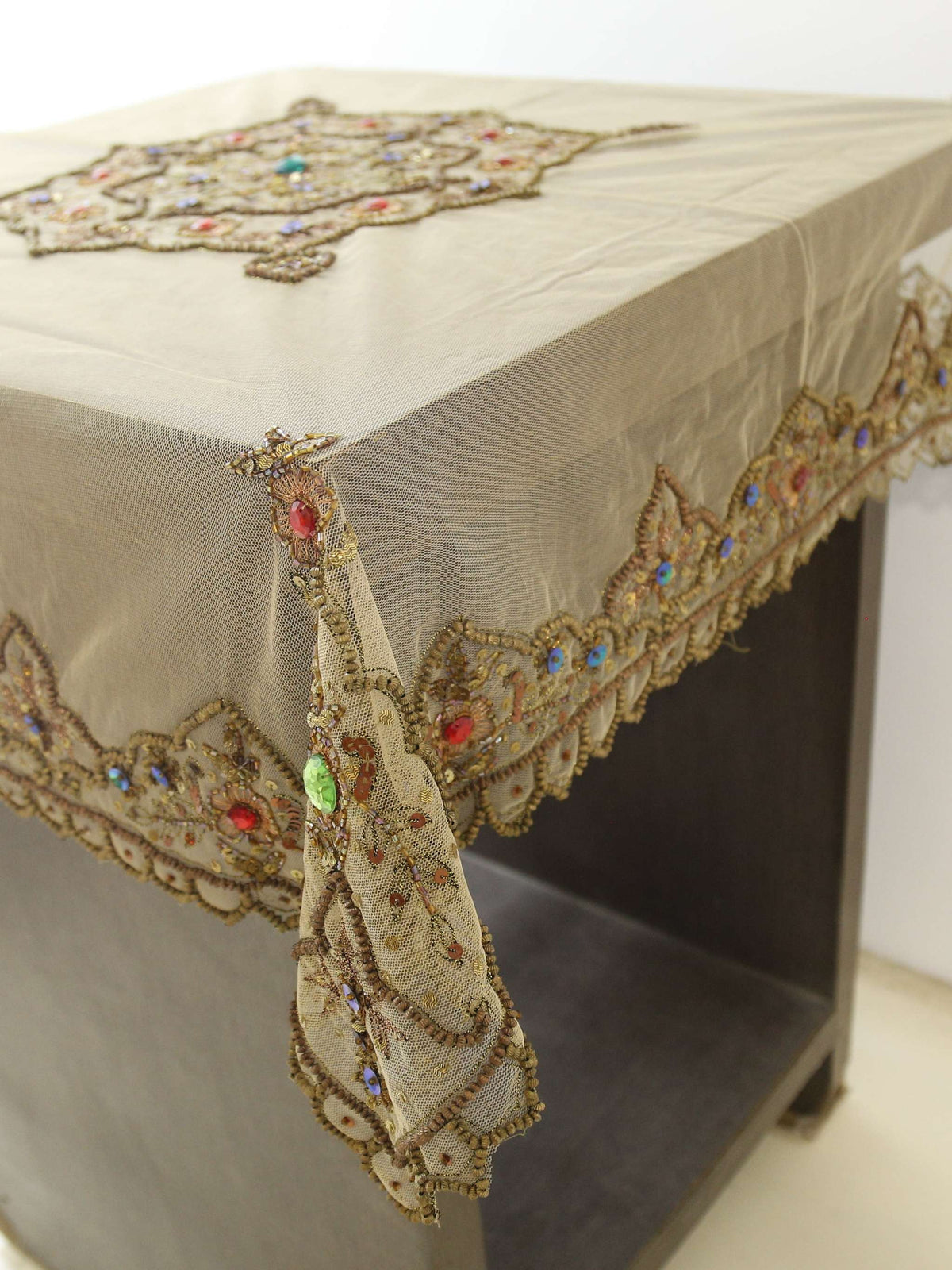 Beige net table cover with a hexagonal gold-beaded floral medallion embroidered at the centre, accented with red and turquoise stones, and a matching beaded scalloped border