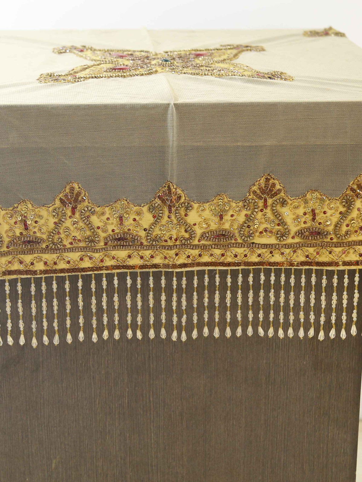 Beige net table cover with a star-shaped gold-beaded medallion embroidered at the centre, accented with red and turquoise stones, edged with a beaded fringe border