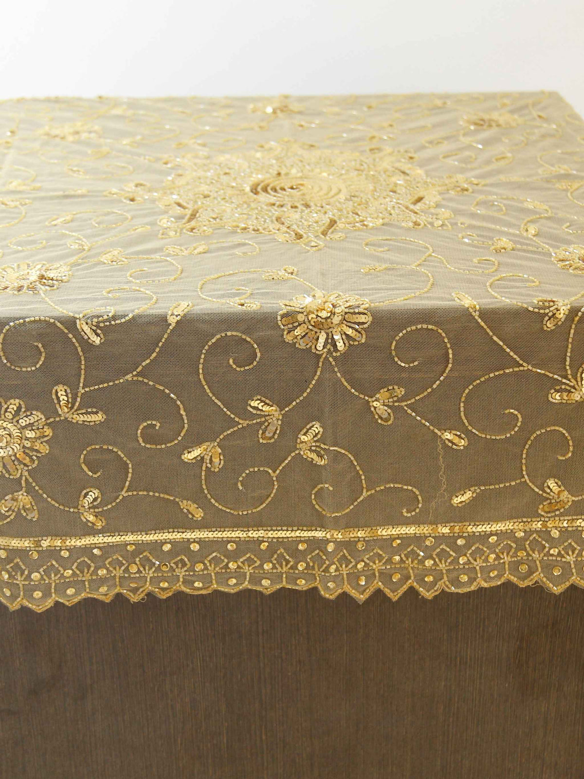 Beige net table cover with a gold sequinned and embroidered floral medallion at the centre and a scalloped lace border