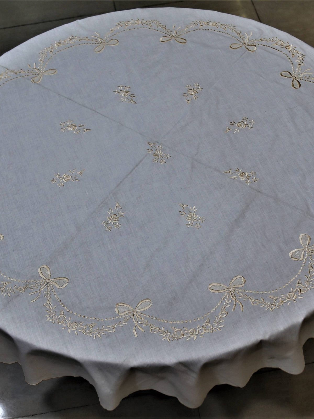 Round ivory table cover with a cream embroidered bow-and-garland pattern and a scalloped edge