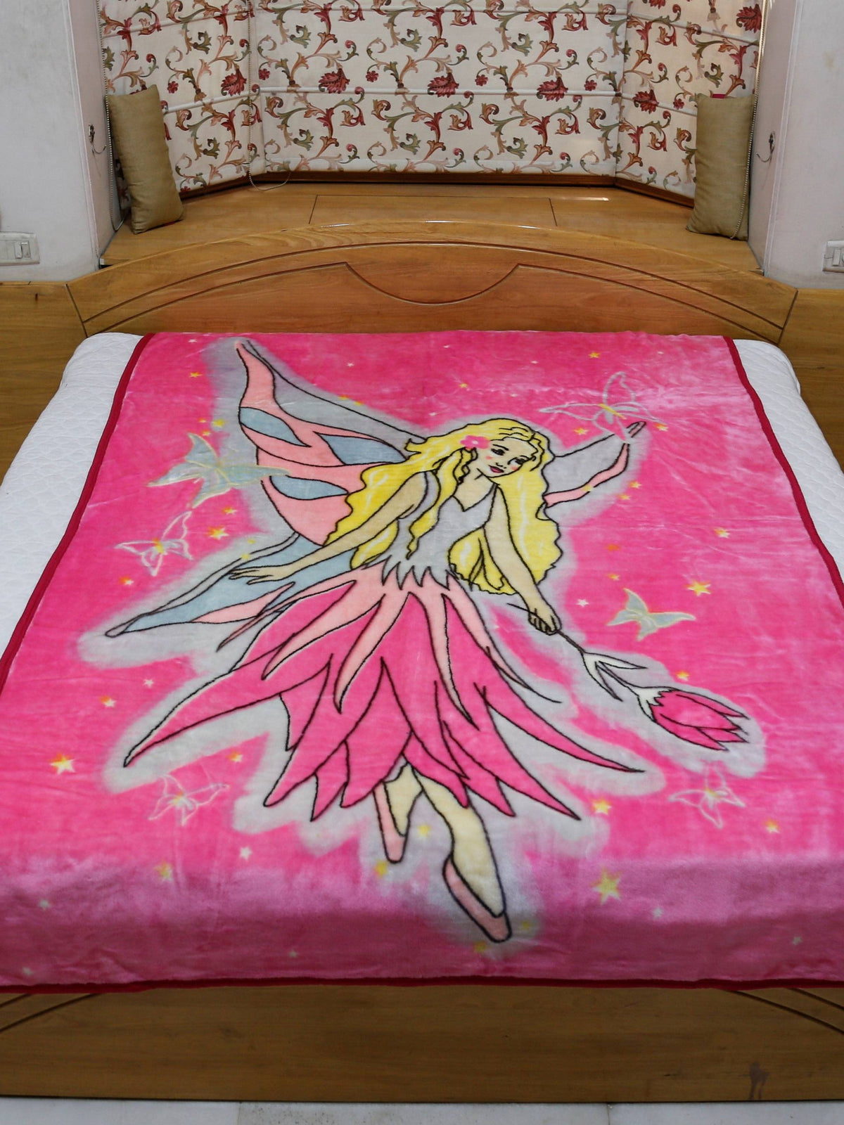 Hot-pink blanket with a printed fairy angel illustration in a flowing pink dress with wings, stars, and butterflies, folded at the foot of a bed