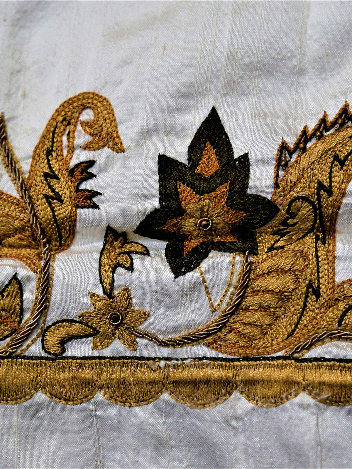 White table cover with a gold-brown embroidered paisley-and-vine corner border and scattered small leaf motifs