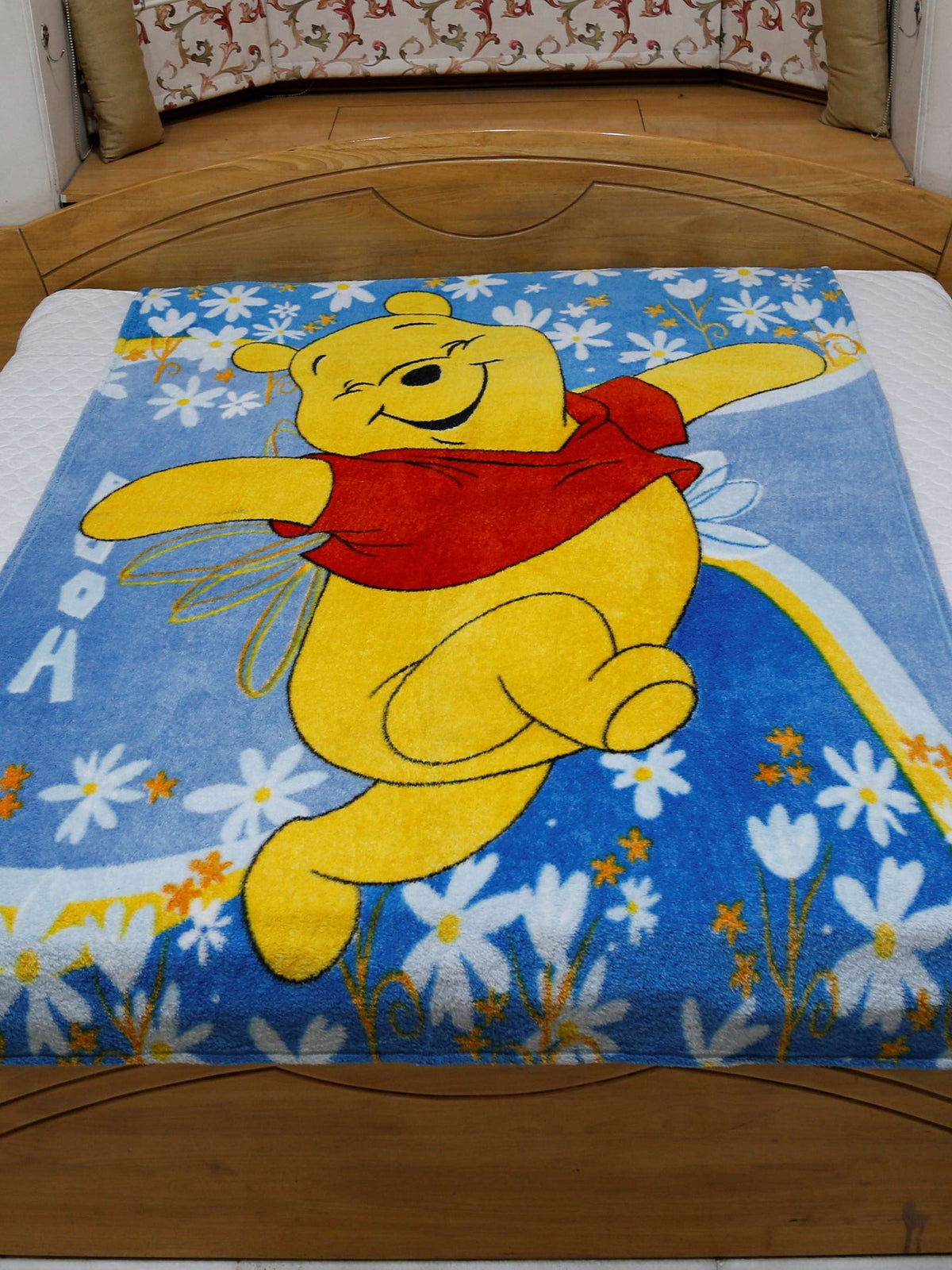 Blue plush blanket with a printed Winnie the Pooh design over a daisy-print border, spread flat over a bed