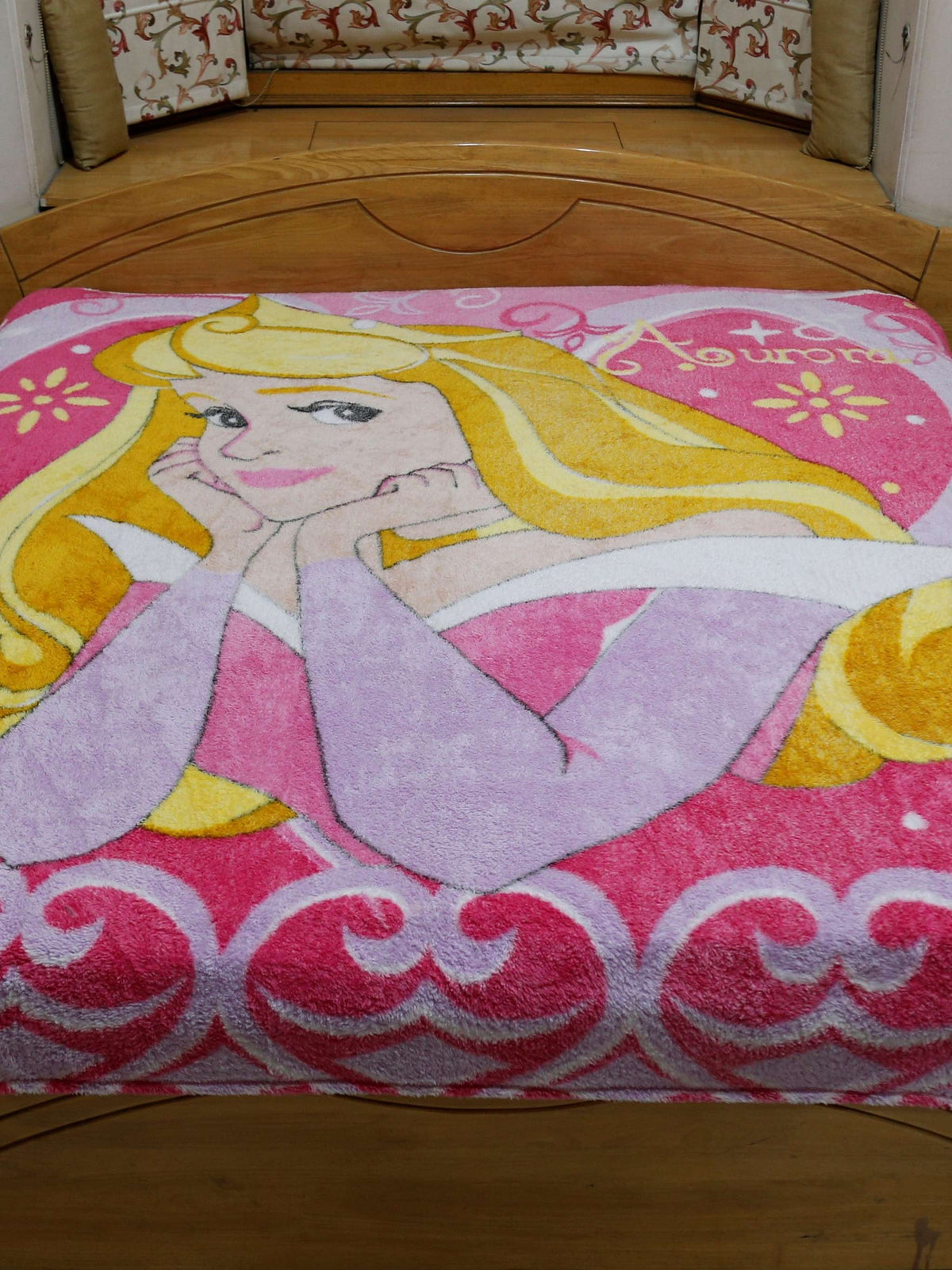 Pink and lilac blanket with a printed Disney Princess Aurora design and a scroll-heart border, spread flat over a bed