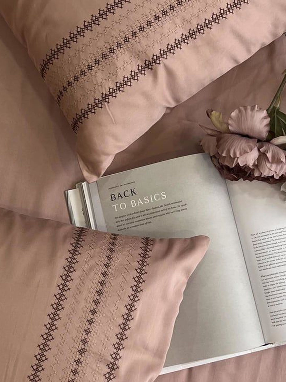 Blush-pink pillow covers with a brown cross-stitch striped embroidered band, styled on a bed with an open book and dried flowers