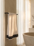 White cotton bath towel with black contrast woven border folded on a wooden bathroom shelf