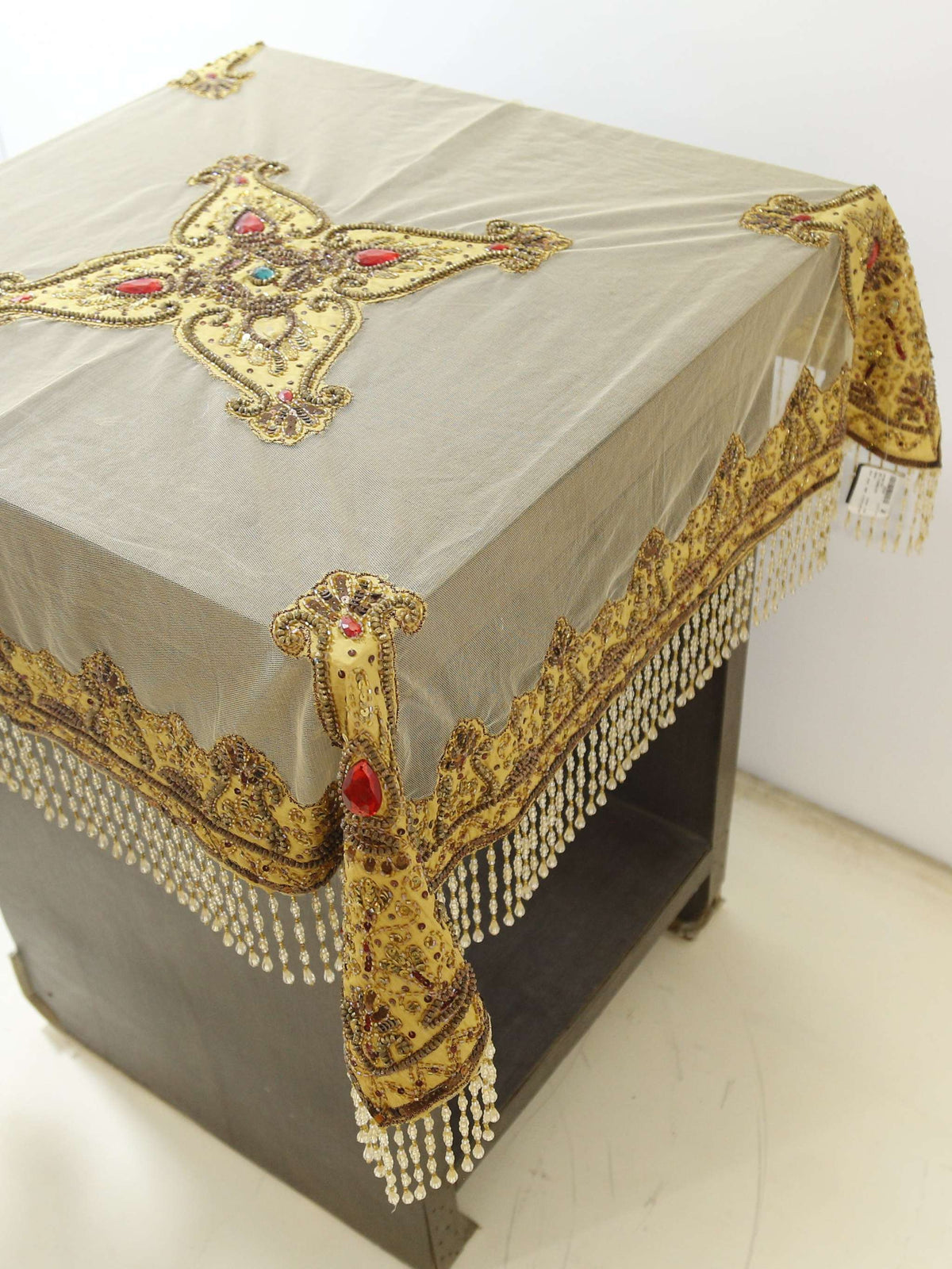 Beige net table cover with a star-shaped gold-beaded medallion embroidered at the centre, accented with red and turquoise stones, edged with a beaded fringe border