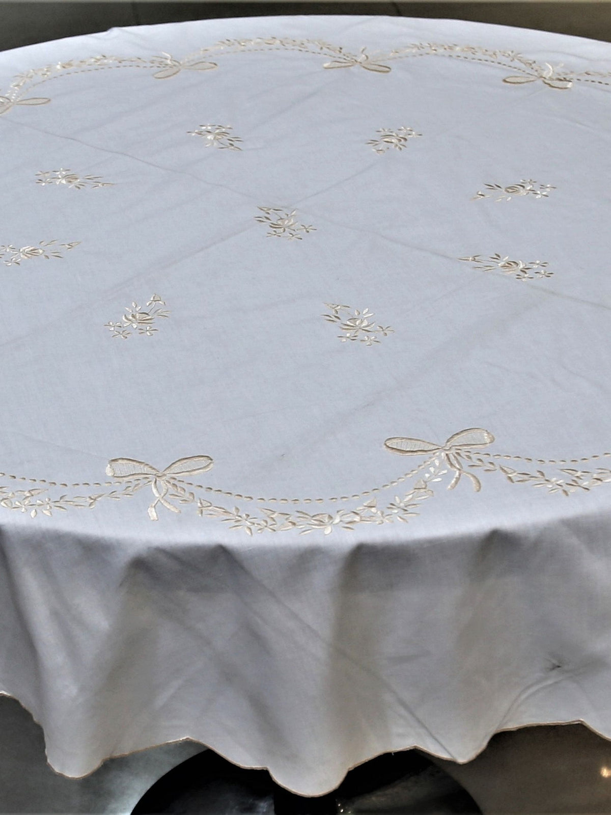 Round ivory table cover with a cream embroidered bow-and-garland pattern and a scalloped edge