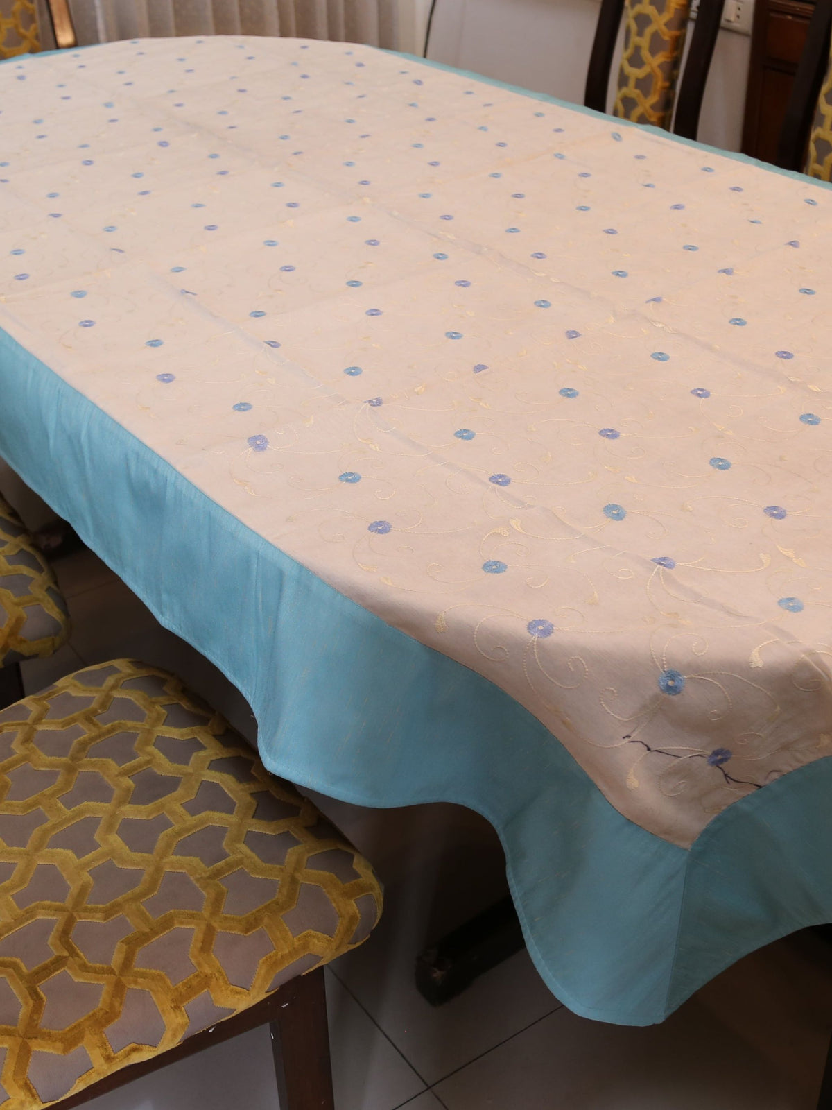 White oval table cover with small blue embroidered flower-and-vine dots and a solid aqua-teal border