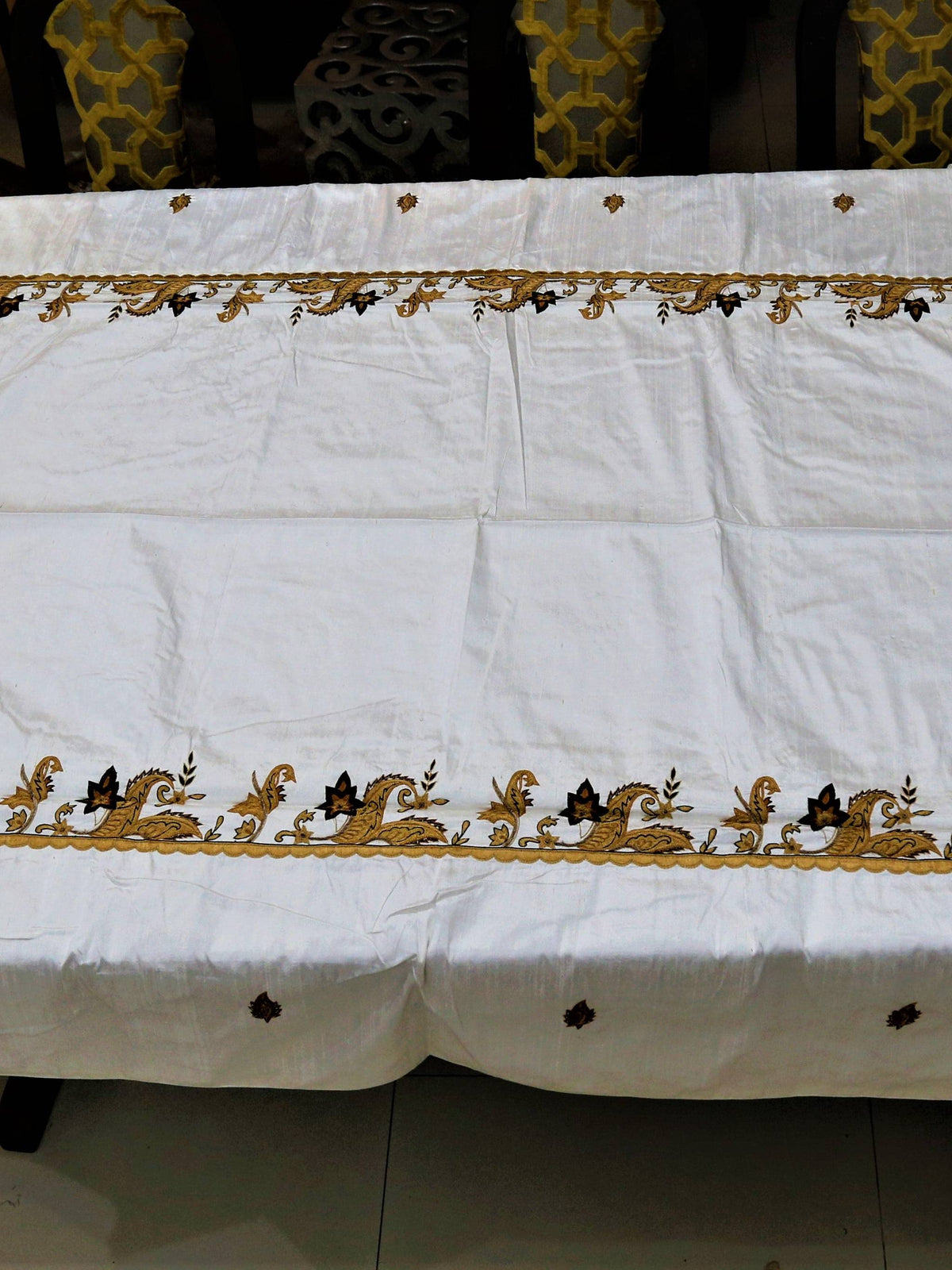 White table cover with a gold-brown embroidered paisley-and-vine corner border and scattered small leaf motifs