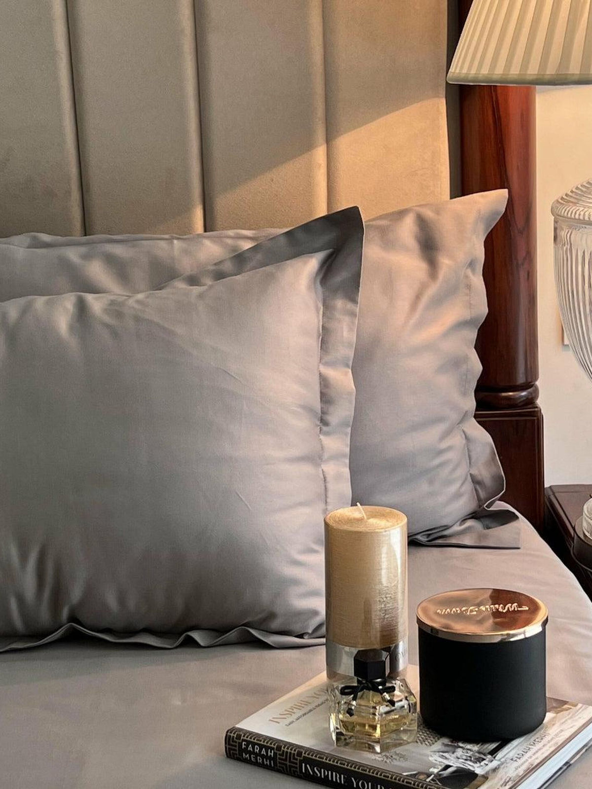 Close-up of grey satin pillow covers on a bed with a scented candle and book on the nightstand