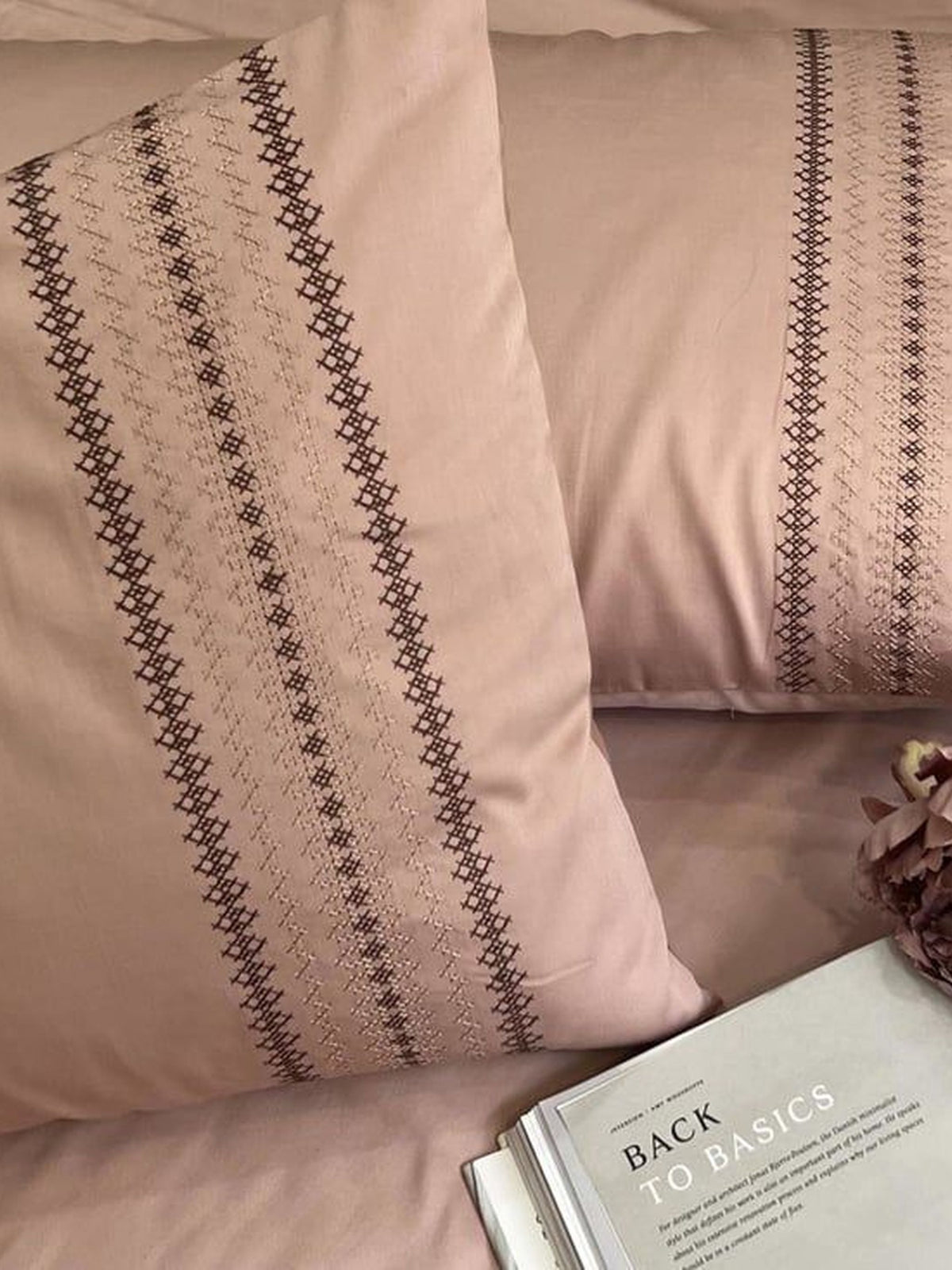 Close-up of blush-pink pillow covers with a brown cross-stitch striped embroidered band, styled on a bed with an open book
