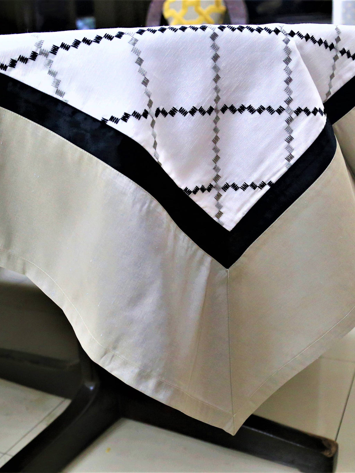 White table cover with a black and grey diamond-lattice embroidered pattern and a plain beige-tan underskirt with a black edge trim