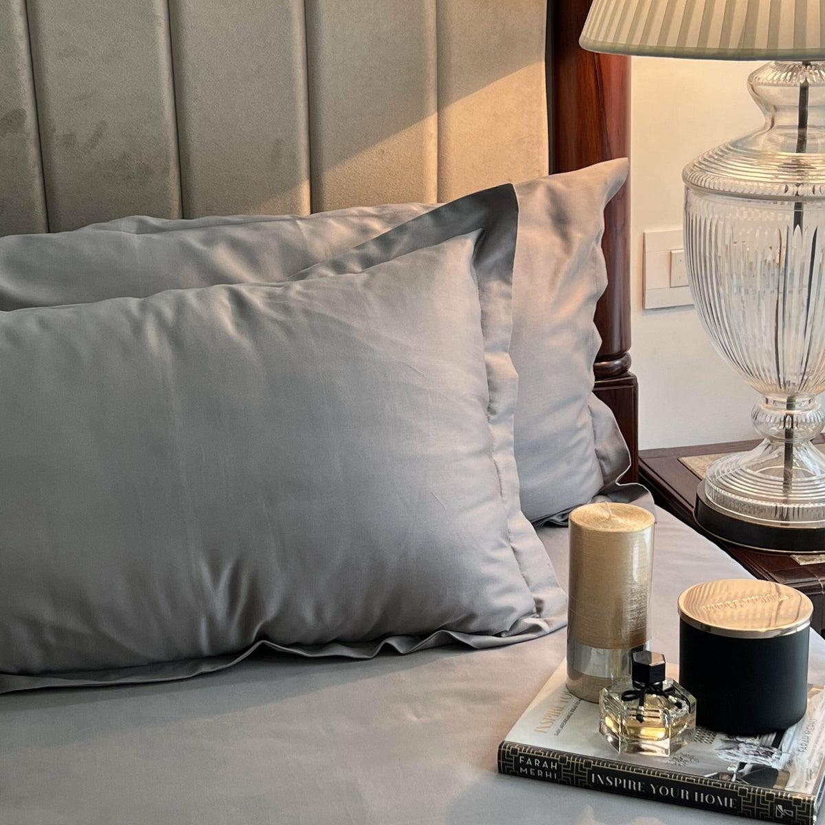 Grey satin pillow covers styled on a bed with a scented candle, perfume bottle, and book on the nightstand