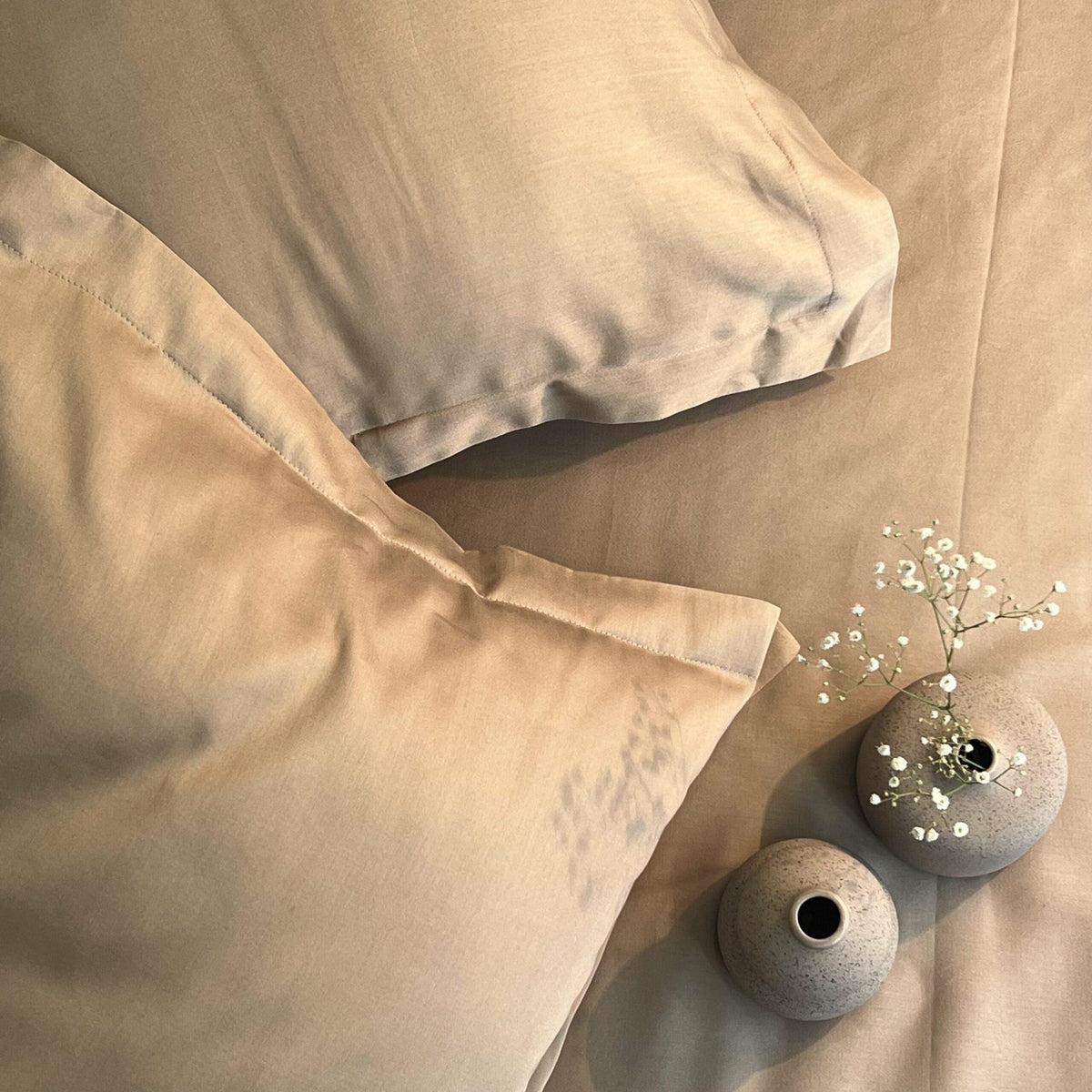 Taupe-sand satin bedsheet and pillow covers styled on a bed with a small vase of blue flowers and baby's breath
