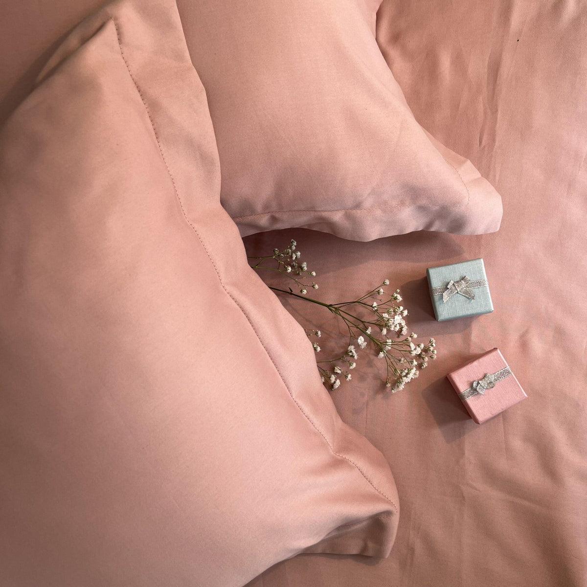 Overhead close-up of dusty rose-pink satin pillow covers on a bed with dried baby's breath flowers and gift boxes