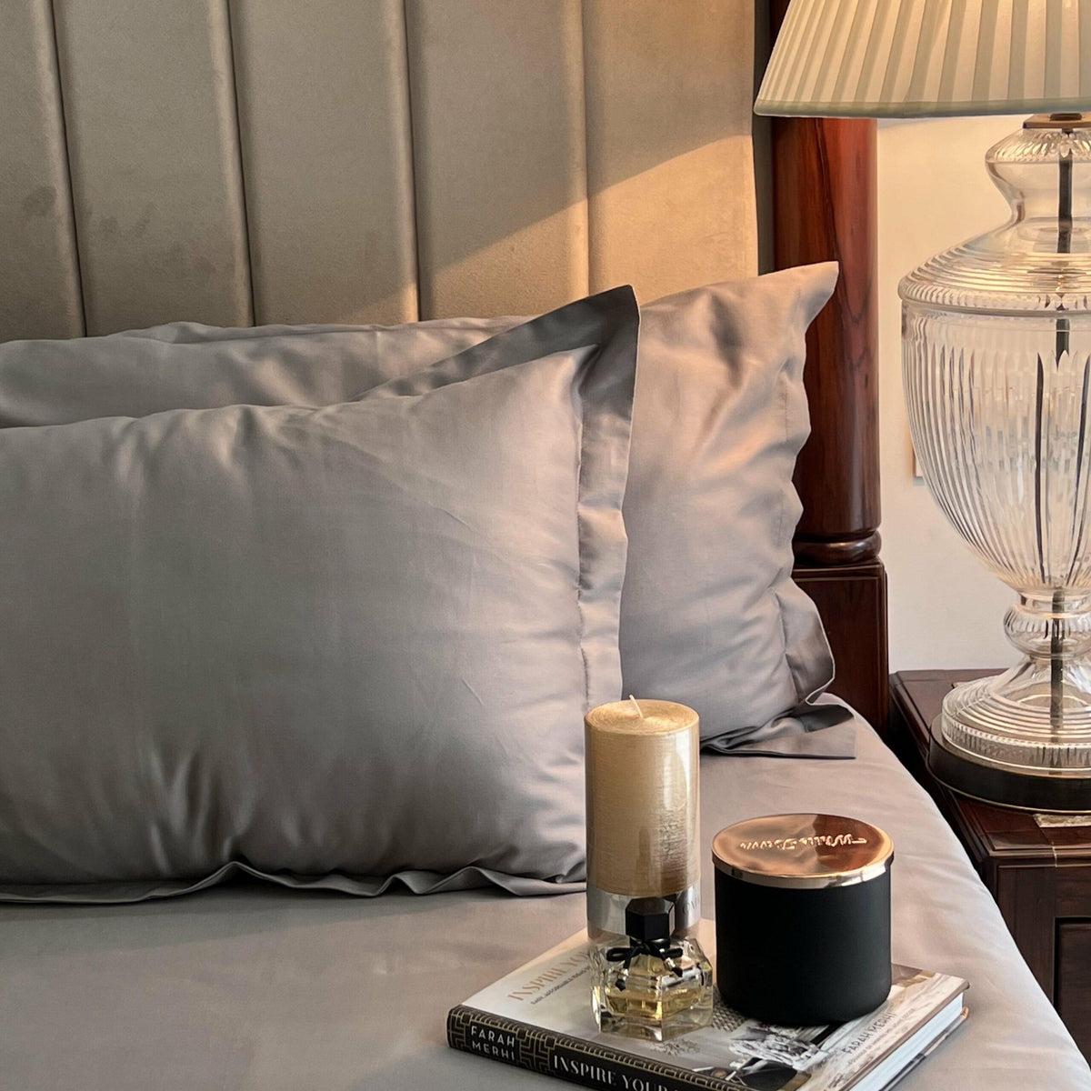 Close-up of grey satin pillow covers on a bed with a scented candle and book on the nightstand