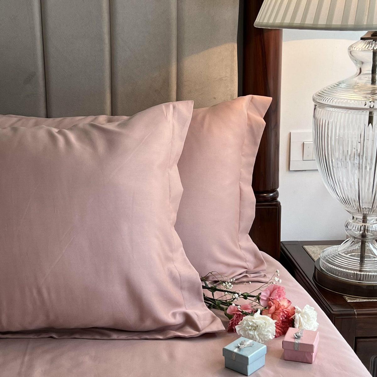 Dusty rose-pink satin pillow covers styled on a bed with dried baby's breath flowers, fresh carnations, and small gift boxes