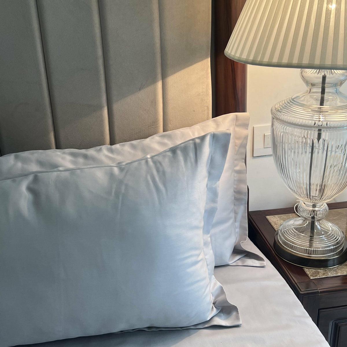 Pale blue-grey satin pillow covers styled on a bed beside a pleated lamp and wooden bedpost