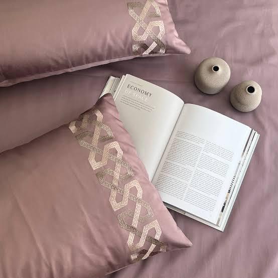 Close-up of dusty mauve-pink pillow covers with a cream trellis-braid embroidered band beside an open book