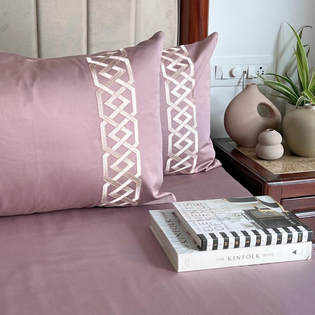 Dusty mauve-pink bedsheet and pillow covers with a cream trellis-braid embroidered band, styled on a bed with a stack of books