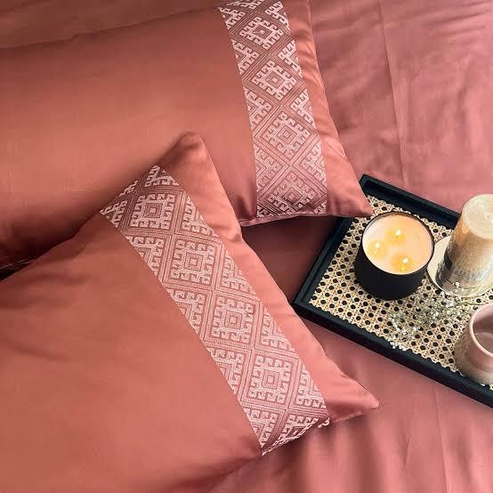 Terracotta bedsheet and pillow covers with a beige diamond-lattice embroidered band, styled on a bed with a lit candle on a cane tray