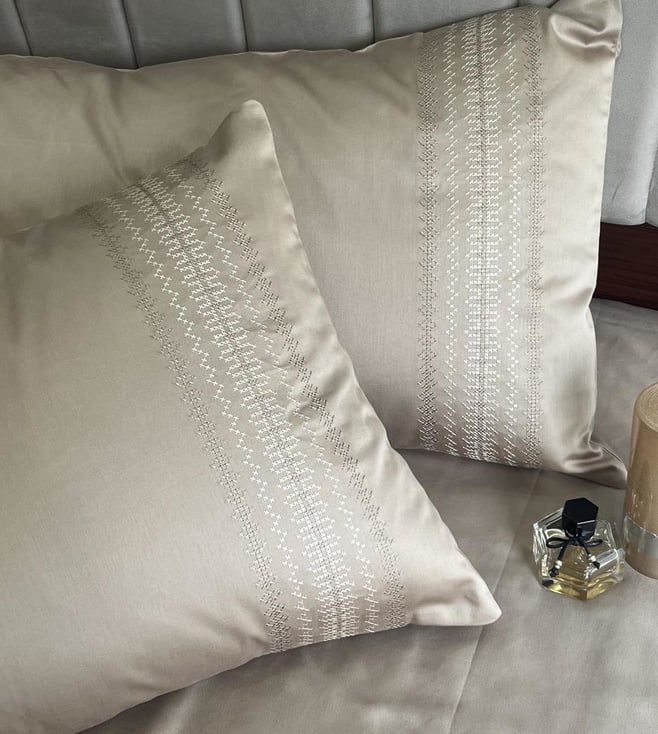 Beige satin pillow covers with a dotted vertical-stripe embroidered band, styled on a bed with a perfume bottle