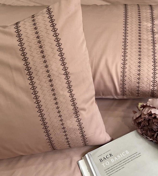 Close-up of blush-pink pillow covers with a brown cross-stitch striped embroidered band, styled on a bed with an open book
