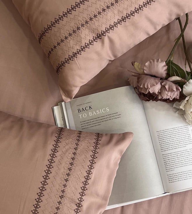 Blush-pink pillow covers with a brown cross-stitch striped embroidered band, styled on a bed with an open book and dried flowers