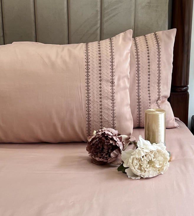 Blush-pink bedsheet and pillow covers with a brown cross-stitch striped embroidered band, styled on a bed with a candle and dried flowers