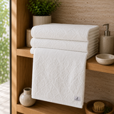 White cotton bath towel with an all-over floral damask pattern folded on a wooden bathroom shelf.