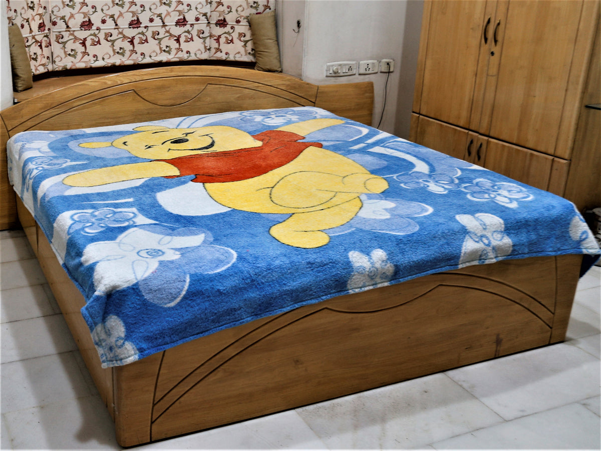 Blue plush blanket with a printed Winnie the Pooh design and white flowers, draped over a bed