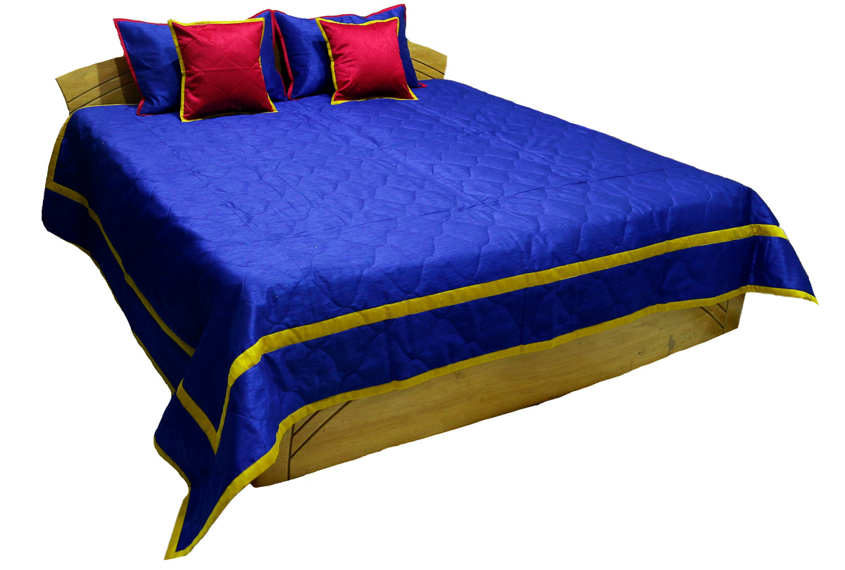 Royal blue quilted bedcover with gold piped border and crimson cushions, angled view